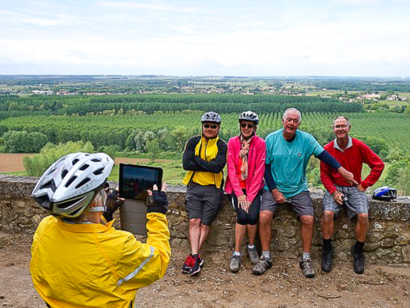 Woman in helmet takes photos of four people sitting on a stone wall with her tablet. In the background are woods and bushes.